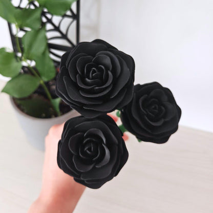 Hand holding a bouquet of three black artificial roses with green stems against a softly blurred plant background.