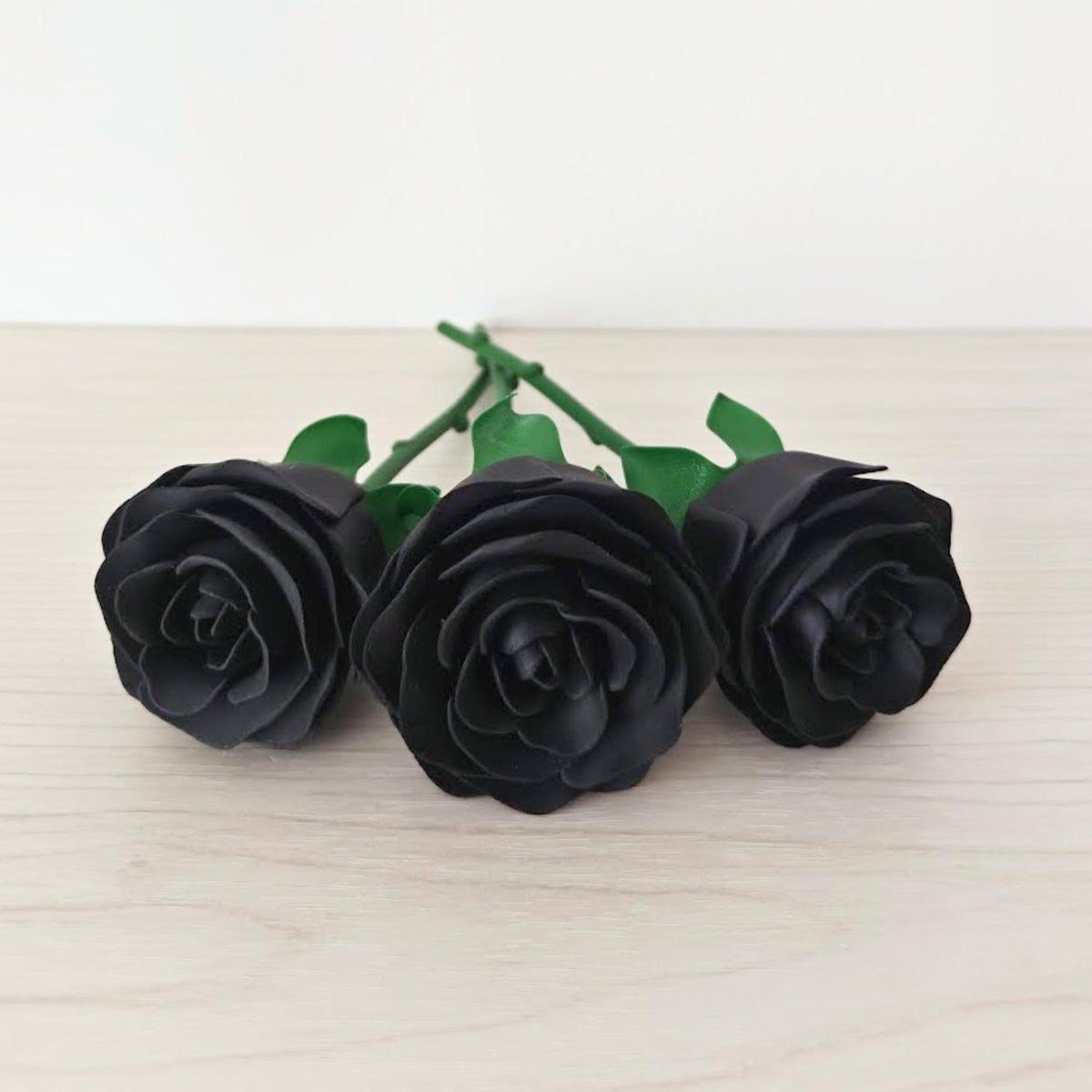 Close-up of three black artificial roses lying side by side on a pale wood surface.