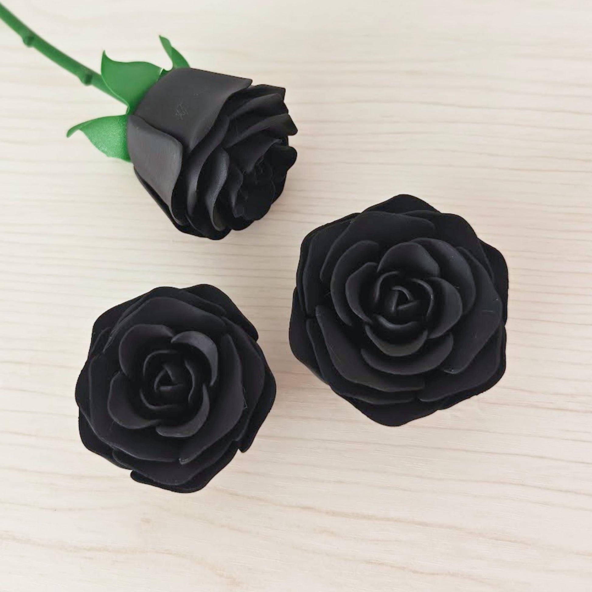 One black rose attached to its green stem beside two detached black rose heads on a light wood surface.