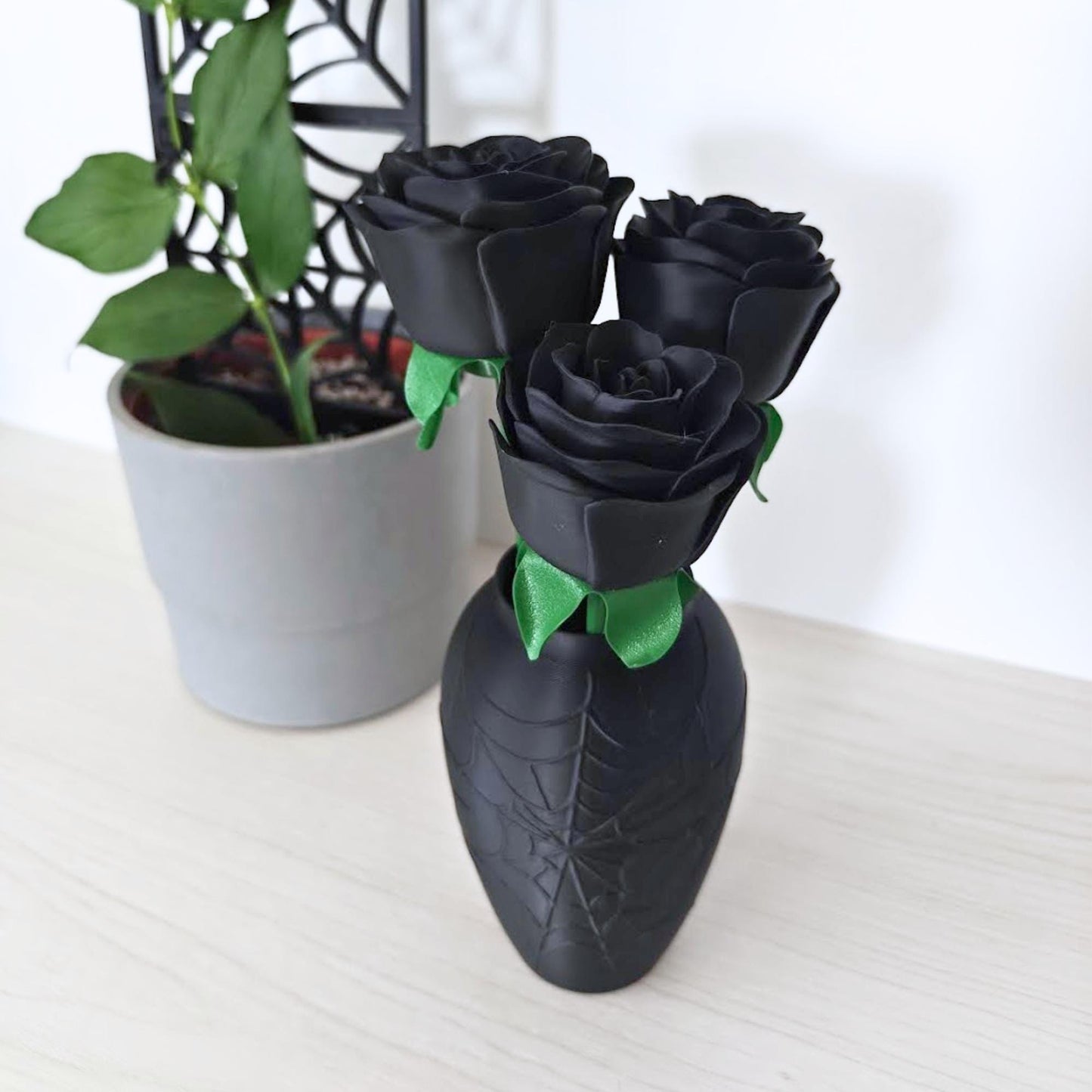 Angled view of the black rose bouquet in its spiderweb vase beside a green houseplant.