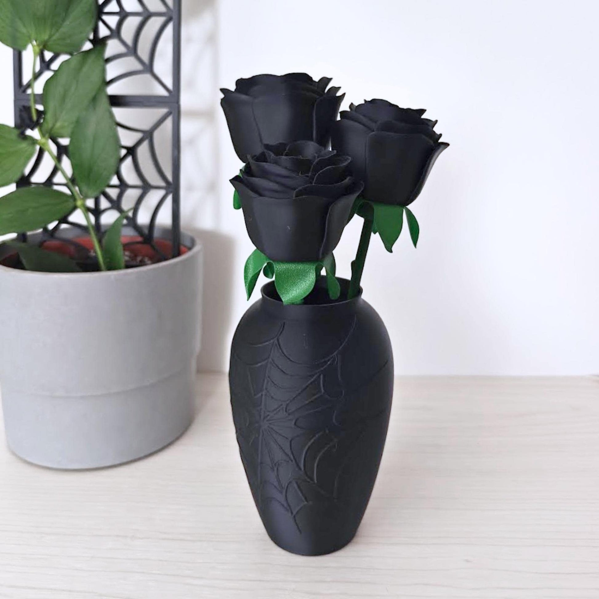 Three matte black faux roses with green stems displayed in a black spiderweb vase beside a grey plant pot.