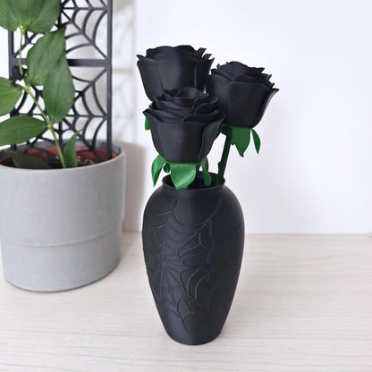 Three matte black faux roses with green stems displayed in a black spiderweb vase beside a grey plant pot.