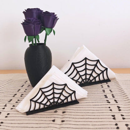 Two black spiderweb napkin holders with folded white napkins displayed beside a black vase with dark purple roses for gothic Halloween table decor.