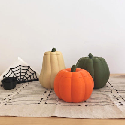 Three lidded pumpkin storage containers in different shapes and colors displayed with black spiderweb Halloween decorations.