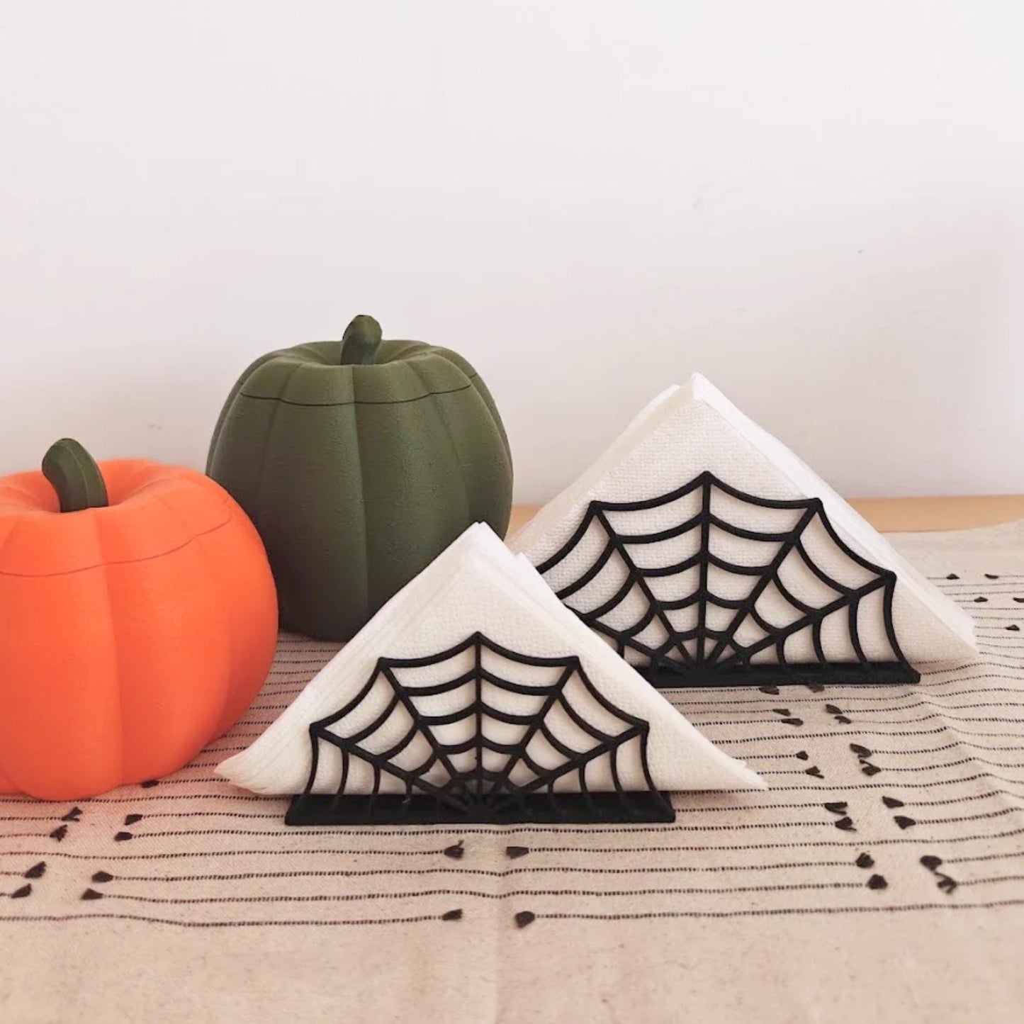 Small and large black spiderweb napkin holders with folded white napkins displayed with orange and dark green pumpkin decorations for an autumn Halloween table setting.
