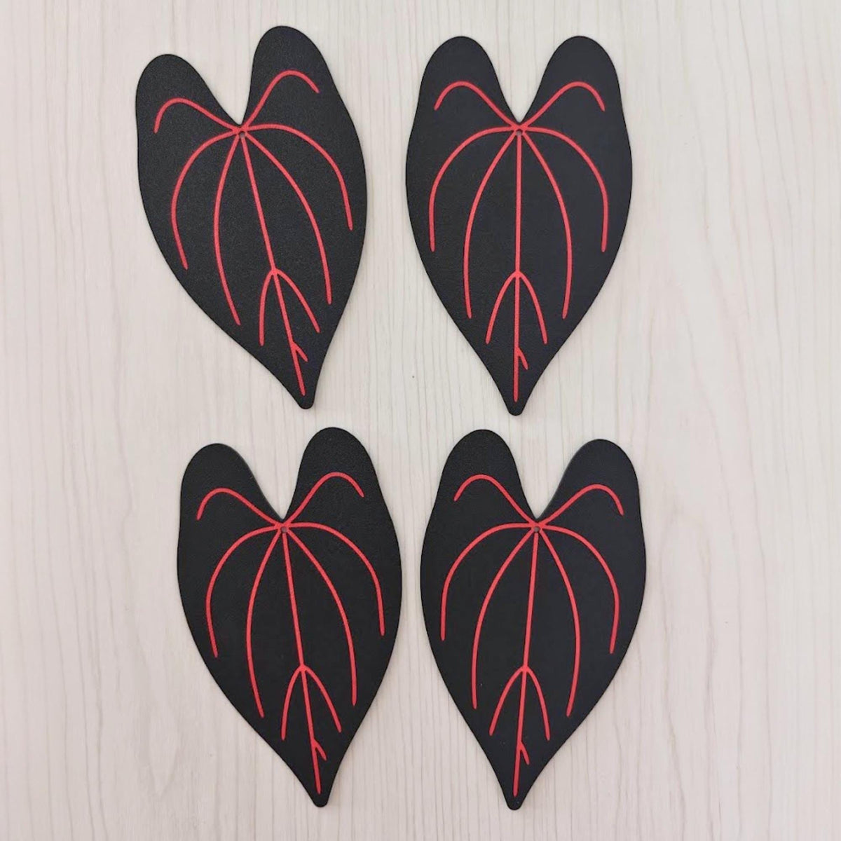 Four black Anthurium Queen of Hearts leaf coasters with bright red veins laid flat on a light wooden surface.
