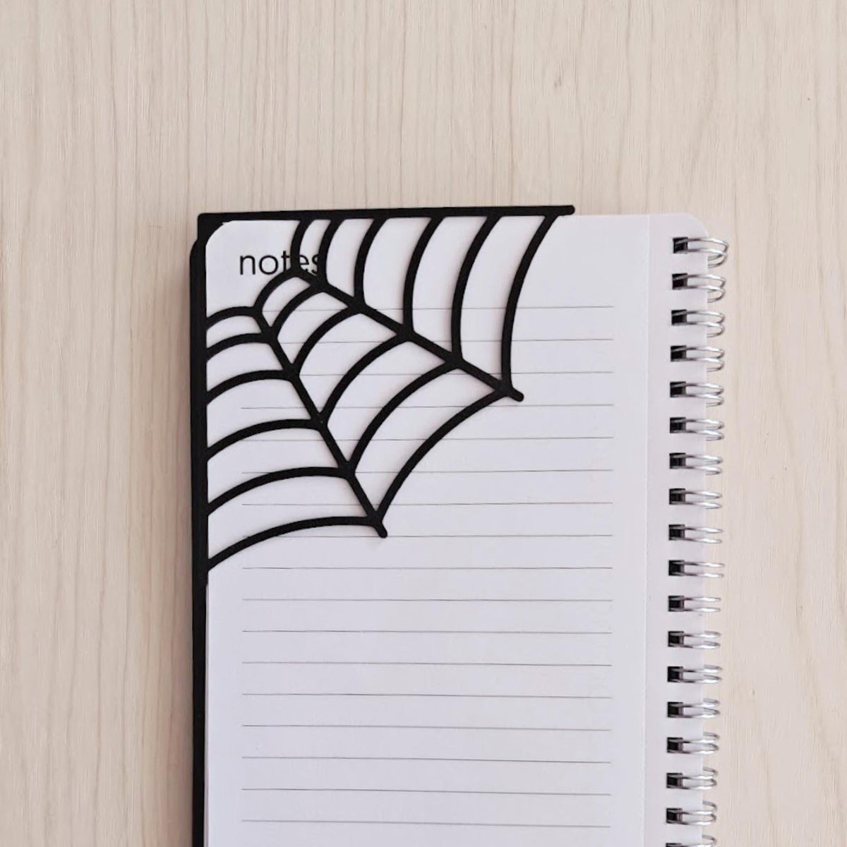 Close-up of a spiderweb corner bookmark attached to the top corner of a spiral notebook page