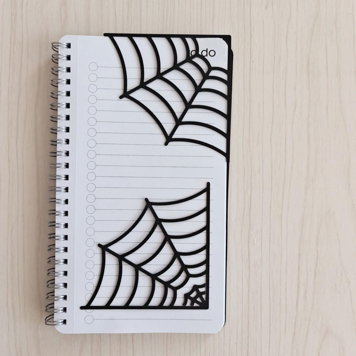 Set of two black spiderweb corner bookmarks displayed on a white spiral notebook