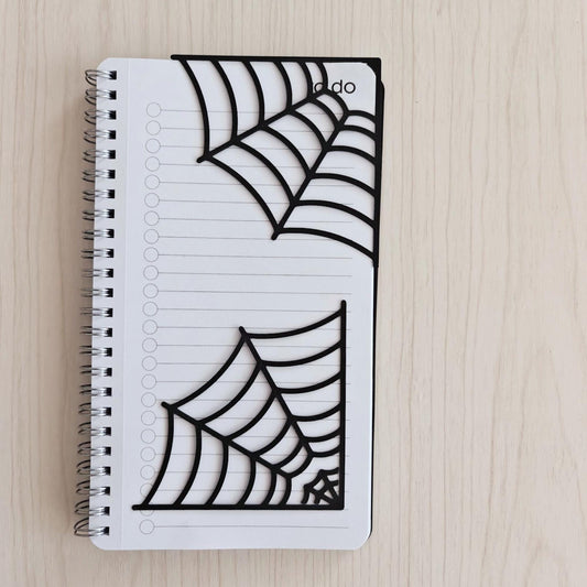 Set of two black spiderweb corner bookmarks displayed on a white spiral notebook