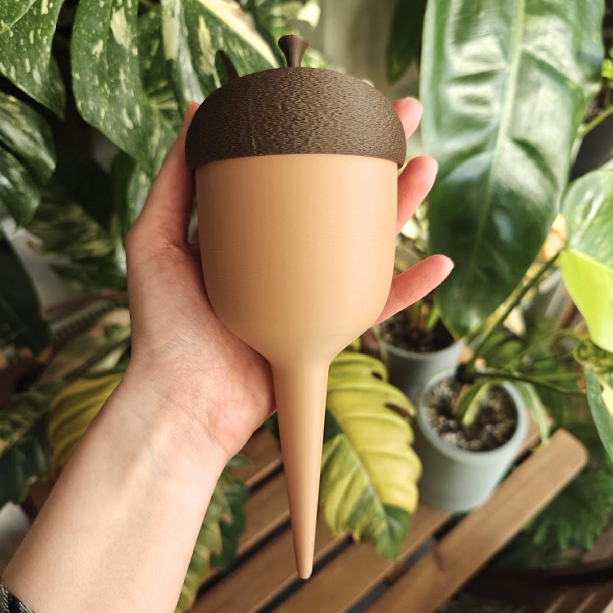 Large acorn-shaped plant watering spike held in one hand in front of green tropical foliage.