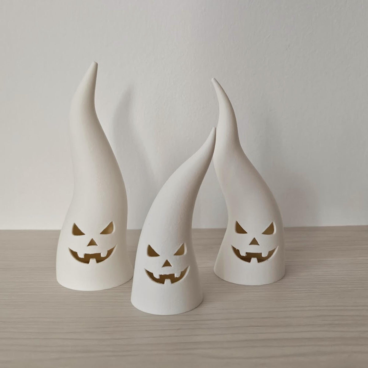 Three white ghost tealight covers with evil faces shown unlit on a light wood surface.