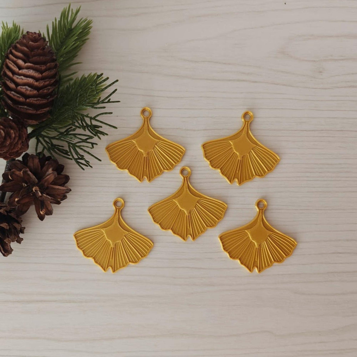 Set of five gold ginkgo leaf Christmas ornaments displayed with pine branches and pinecones.