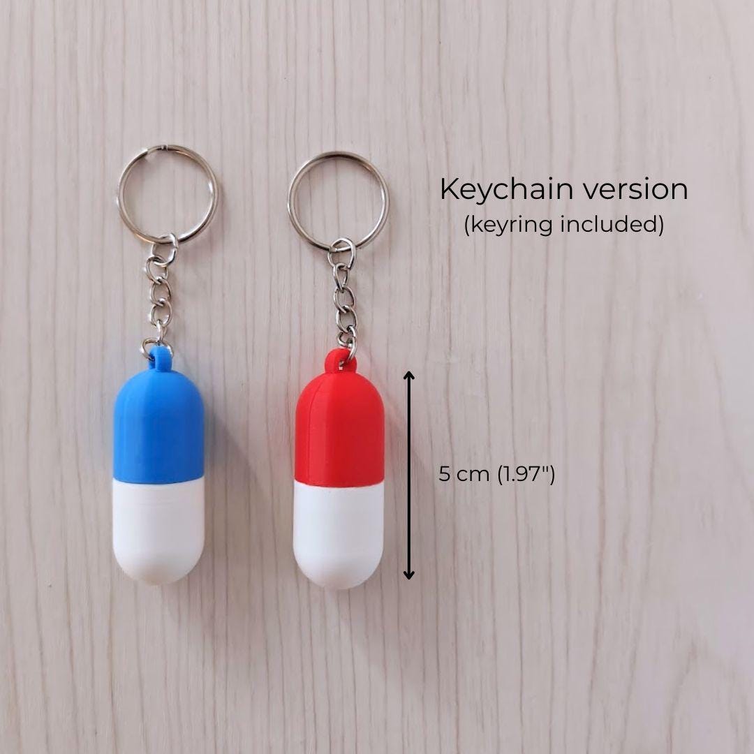 Two capsule-shaped keychain pill containers in blue/white and red/white, shown with keyrings and 5 cm size reference.